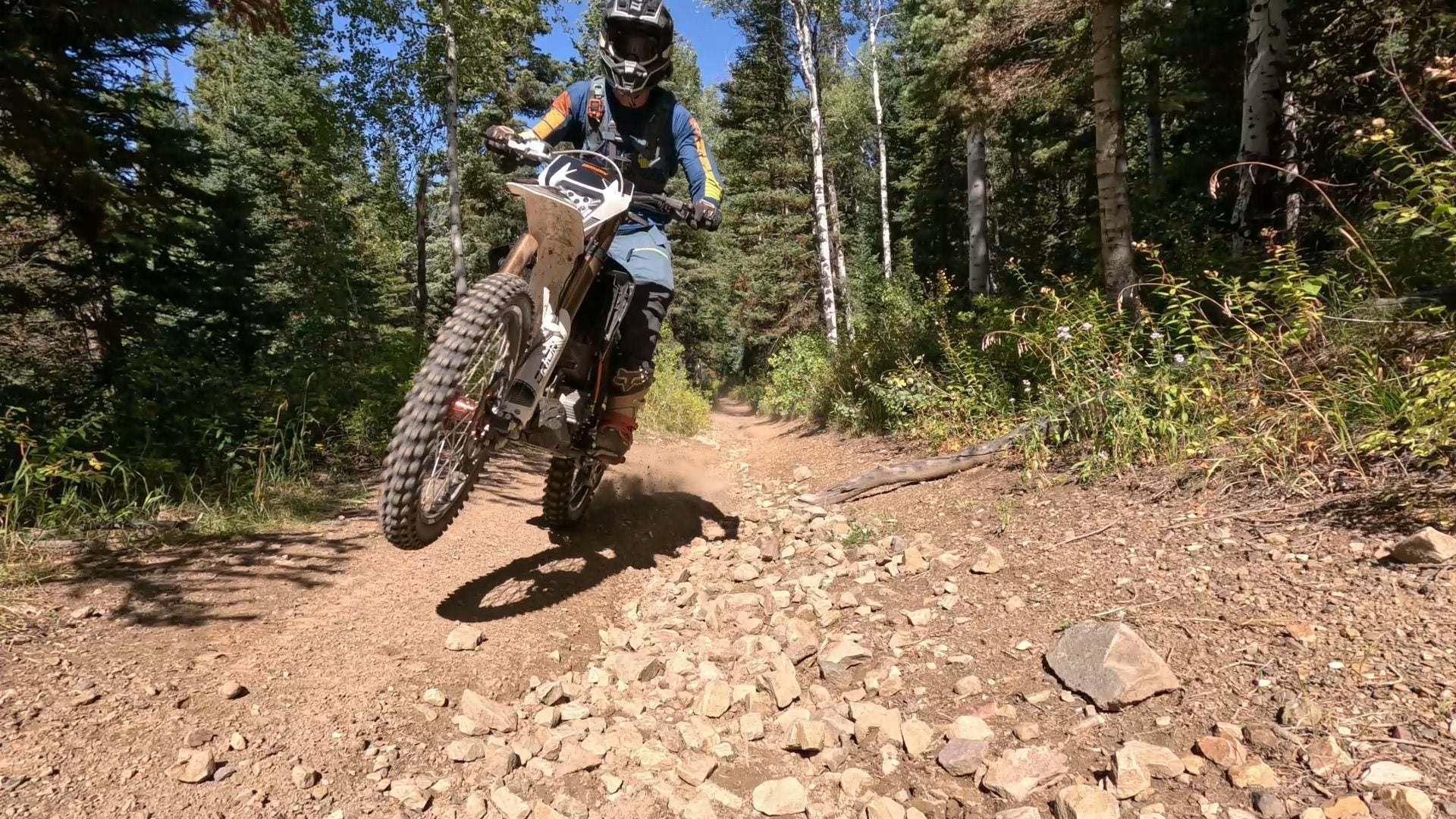 A motocross rider wearing full protective gear navigates a rocky forest trail on an Orion electric dirt bike, lifting the front wheel slightly off the ground.